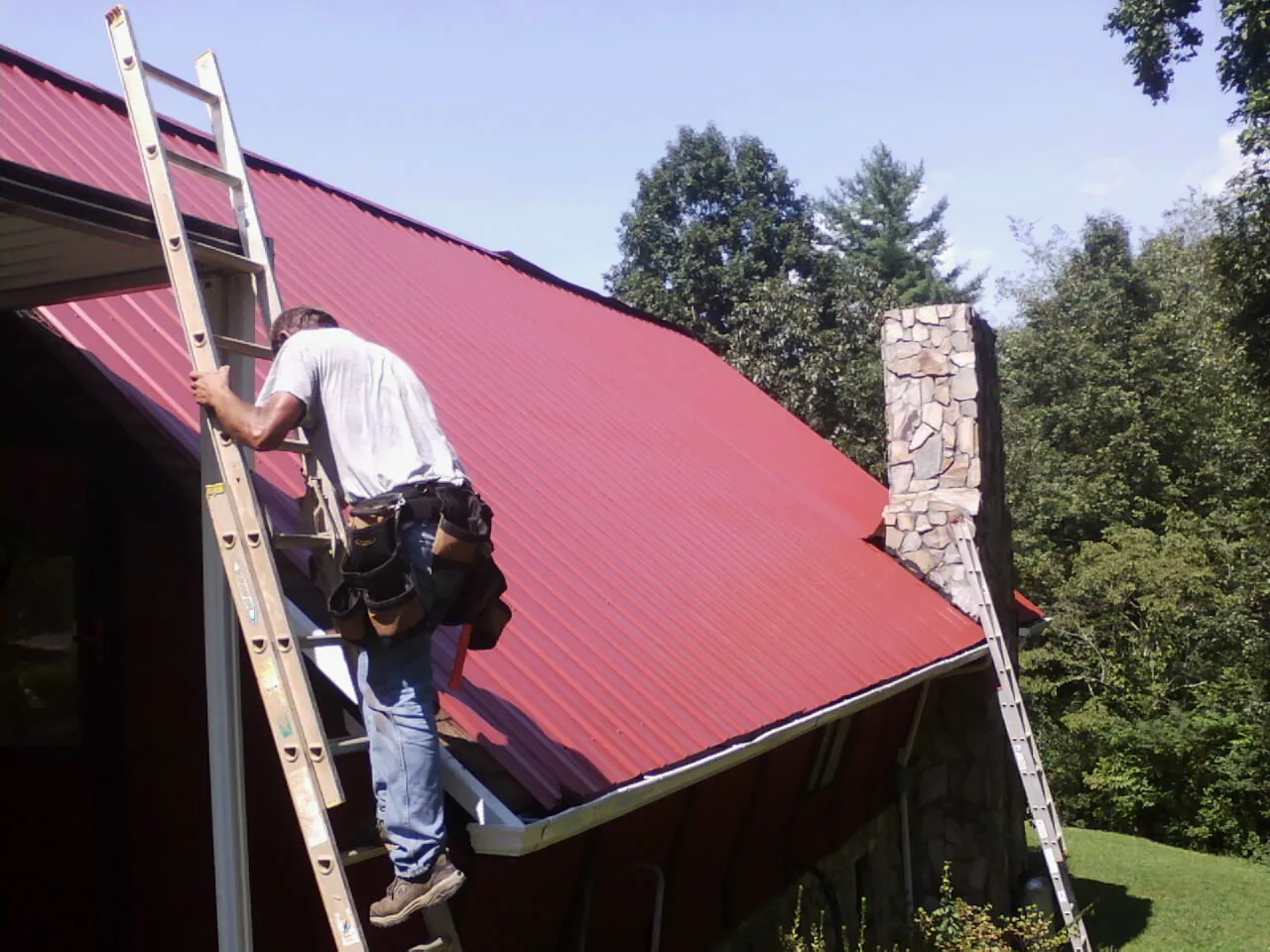 Roofer on a ladder beside a red metal roof and stone chimney — Soffit & Fascia Repair by Roofing Brothers in Hubertus